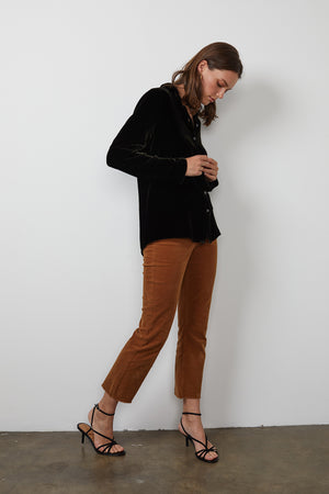 A person wearing the JENSINE SILK VELVET BUTTON-UP SHIRT in black from Velvet by Graham & Spencer, paired with brown pants and black strappy heels, adjusts their top while facing slightly to the right against a plain white background, subtly hinting at the smooth elegance of velvet in their ensemble.
