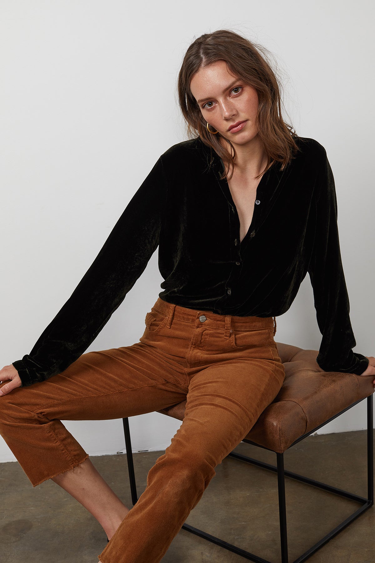 A woman wearing a black JENSINE Silk Velvet Button-Up Shirt by Velvet by Graham & Spencer and brown pants sits on a brown velvet cushioned bench against a plain white wall.-15201374339265