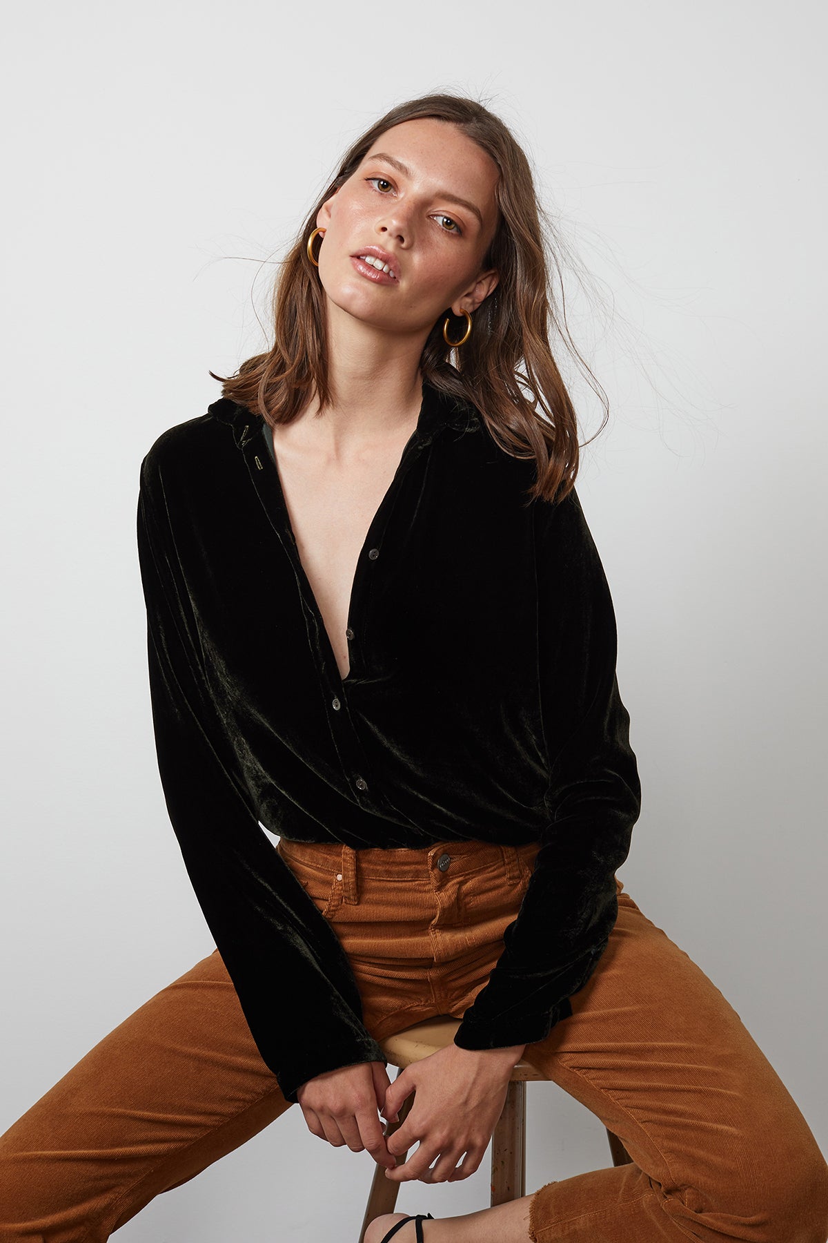 A woman wearing the JENSINE SILK VELVET BUTTON-UP SHIRT by Velvet by Graham & Spencer, paired with brown pants, is sitting on a stool against a plain white background, looking directly at the camera.-15201374142657