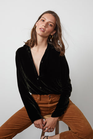 A woman wearing the JENSINE SILK VELVET BUTTON-UP SHIRT by Velvet by Graham & Spencer, paired with brown pants, is sitting on a stool against a plain white background, looking directly at the camera.