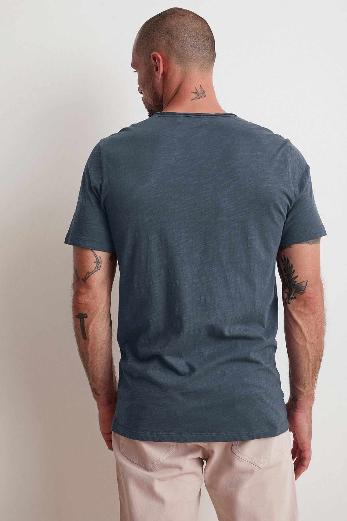 A man with short hair and tattoos on his arms is shown from the back, wearing a plain, dark gray CHAD RAW EDGE COTTON SLUB POCKET TEE by Velvet by Graham & Spencer and light beige pants. The tee features raw edge details, adding a subtle touch against the white background he stands before.-25792954532033