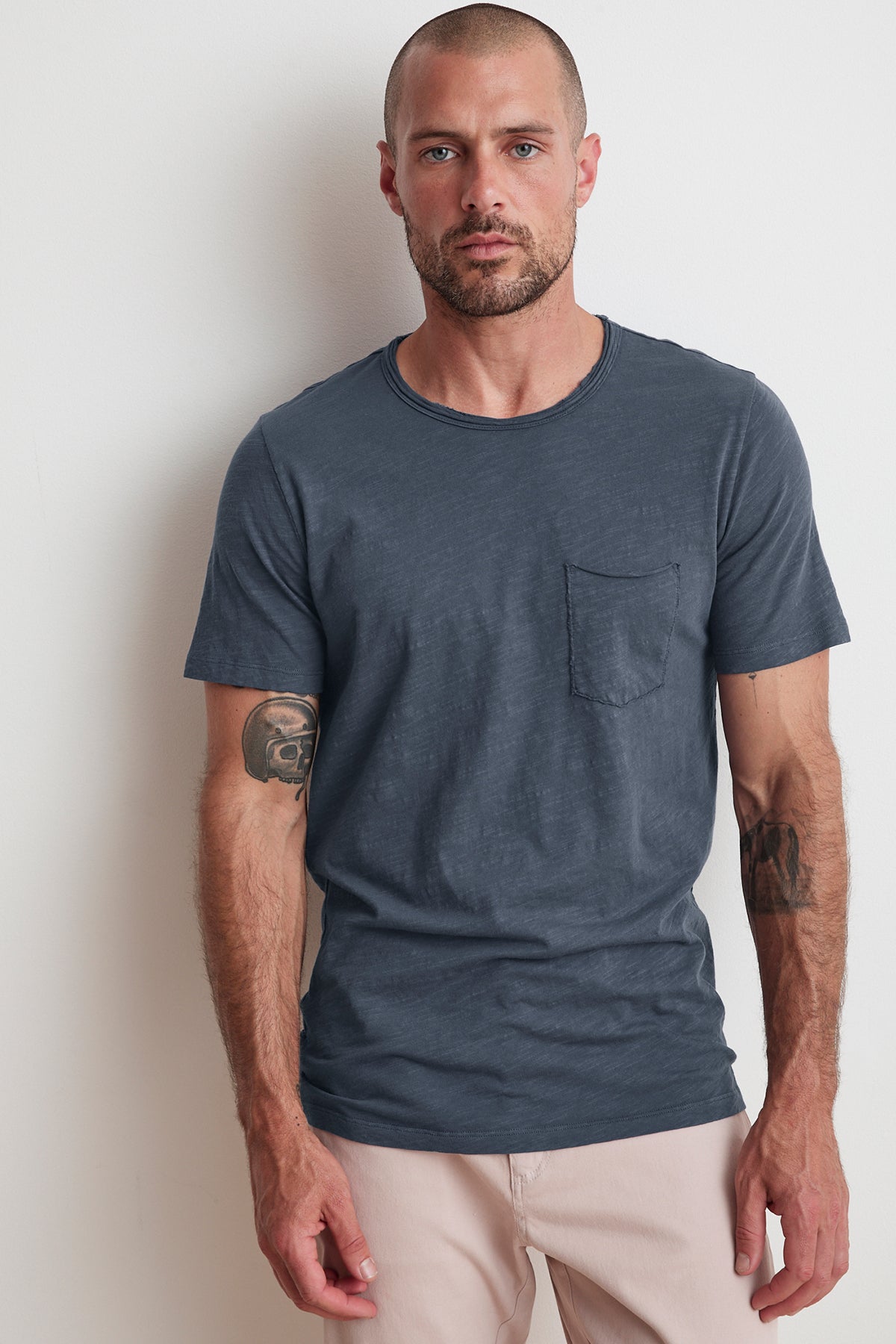 A man with a beard and tattoos on both arms is wearing a Velvet by Graham & Spencer CHAD RAW EDGE COTTON SLUB POCKET TEE, paired with light-colored pants, standing against a plain white wall.