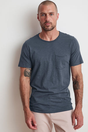 A man with a beard and tattoos on both arms is wearing a Velvet by Graham & Spencer CHAD RAW EDGE COTTON SLUB POCKET TEE, paired with light-colored pants, standing against a plain white wall.