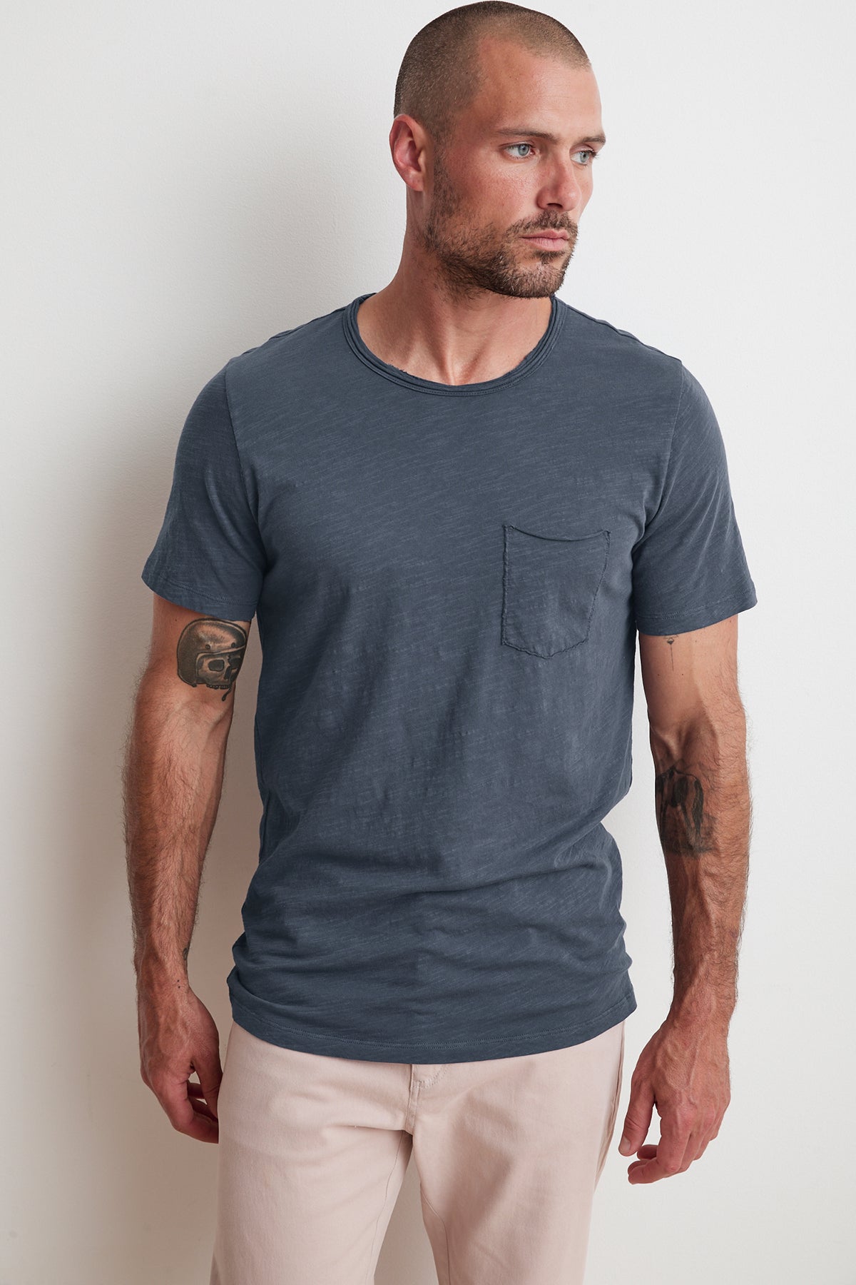 A man with a beard, short hair, and tattoos on his forearms is wearing a Velvet by Graham & Spencer CHAD RAW EDGE COTTON SLUB POCKET TEE with classic crew neckline and light-colored pants, standing against a plain white wall, looking to his right.