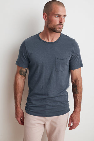 A man with a beard, short hair, and tattoos on his forearms is wearing a Velvet by Graham & Spencer CHAD RAW EDGE COTTON SLUB POCKET TEE with classic crew neckline and light-colored pants, standing against a plain white wall, looking to his right.