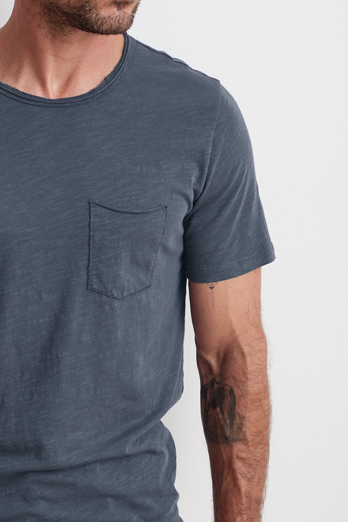 Close-up of a man wearing a faded blue Velvet by Graham & Spencer CHAD RAW EDGE COTTON SLUB POCKET TEE, standing against a white background. His left forearm is visible, showing a tattoo.-25792954597569