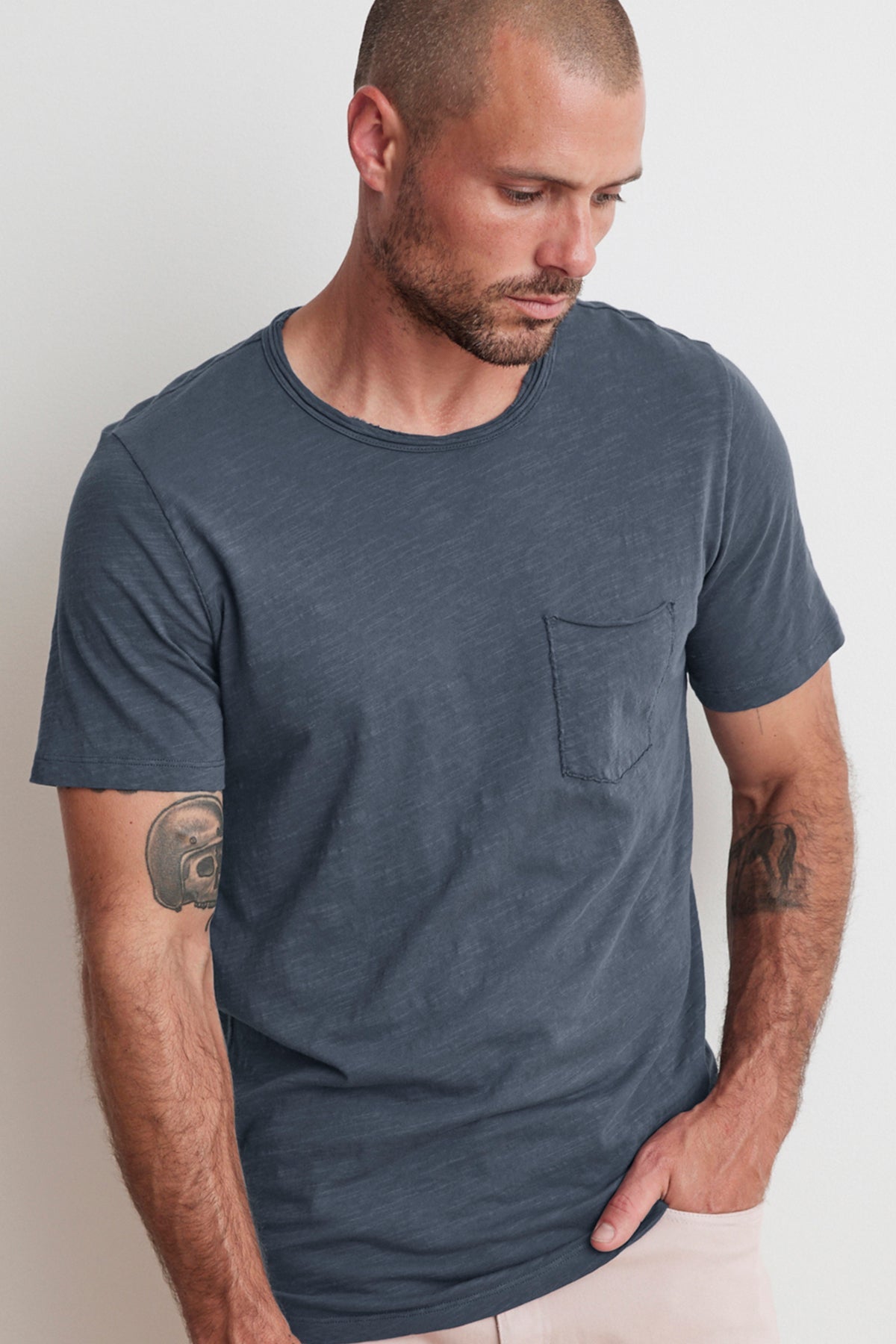 A man with a shaved head and tattoos on his arms is looking down while wearing the CHAD RAW EDGE COTTON SLUB POCKET TEE by Velvet by Graham & Spencer.-25792954499265