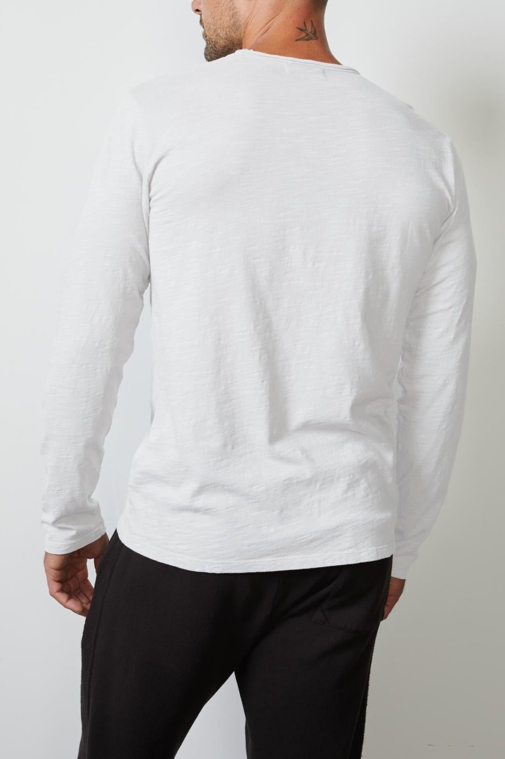 A man with a shaved head is seen from the back, wearing a white long-sleeve SIMEON TEE by Velvet by Graham & Spencer made of soft slub cotton and black pants. He has a small tattoo on the back of his neck.-26146678309057