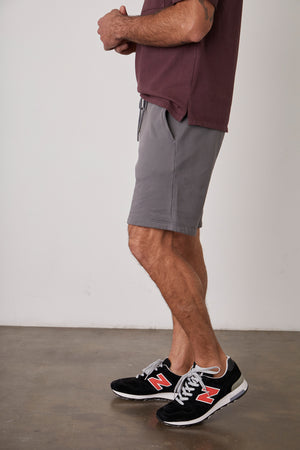 A man wearing Velvet by Graham & Spencer JAXSON DRAWSTRING SHORT and sneakers standing in front of a white wall.