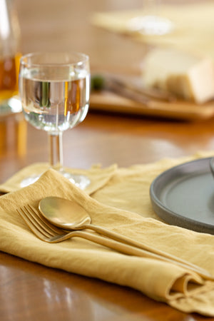 A neatly set dining table with an elegant yellow Jenny Graham Home linen napkin, a spoon, and a glass of water, with blurry plates and a meal in the background.