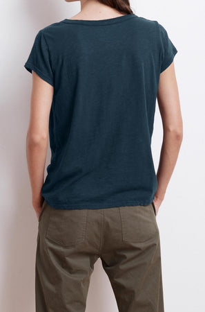 A person is standing with their back to the camera, dressed in everyday wear that includes a dark blue short-sleeved KIRA TEE by Velvet by Graham & Spencer made of cotton slub fabric and olive green pants.