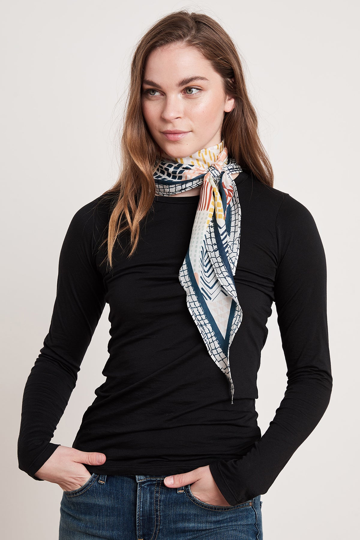 A young woman, dressed in a black long-sleeve shirt and blue jeans, stands against a plain background with her hands in her pockets. Around her neck is the DIAMOND SHAPE SILK SCARF by Velvet by Graham & Spencer, which features a colorful pattern.-6705365286993