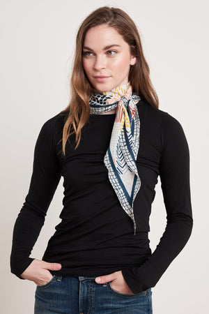 A young woman, dressed in a black long-sleeve shirt and blue jeans, stands against a plain background with her hands in her pockets. Around her neck is the DIAMOND SHAPE SILK SCARF by Velvet by Graham & Spencer, which features a colorful pattern.