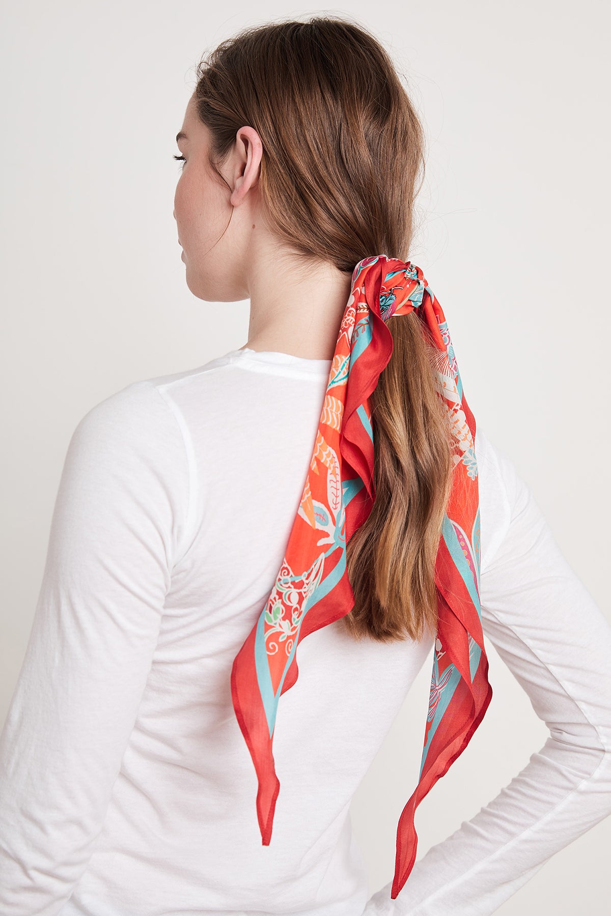 A woman with long brown hair tied in a ponytail, adorned with the Velvet by Graham & Spencer DIAMOND SHAPE SILK SCARF, wearing a white long-sleeve shirt and turned slightly sideways.