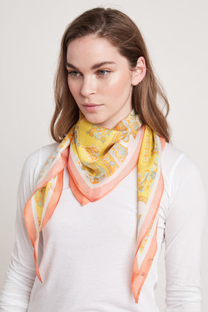 A person wearing a white shirt and a Diamond Shape Silk Scarf by Velvet by Graham & Spencer, featuring a yellow pattern with pink edges, wrapped around their neck.