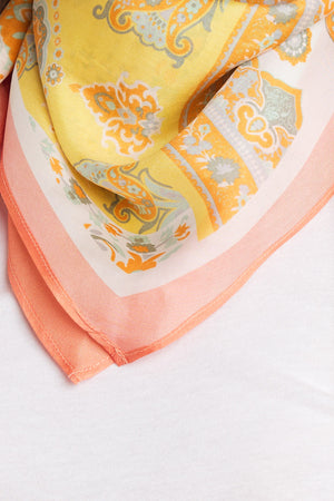 A close-up of the DIAMOND SHAPE SILK SCARF from Velvet by Graham & Spencer, showcasing its vibrant yellow, orange, and green floral patterns, draped over a white fabric background in a diamond silhouette.