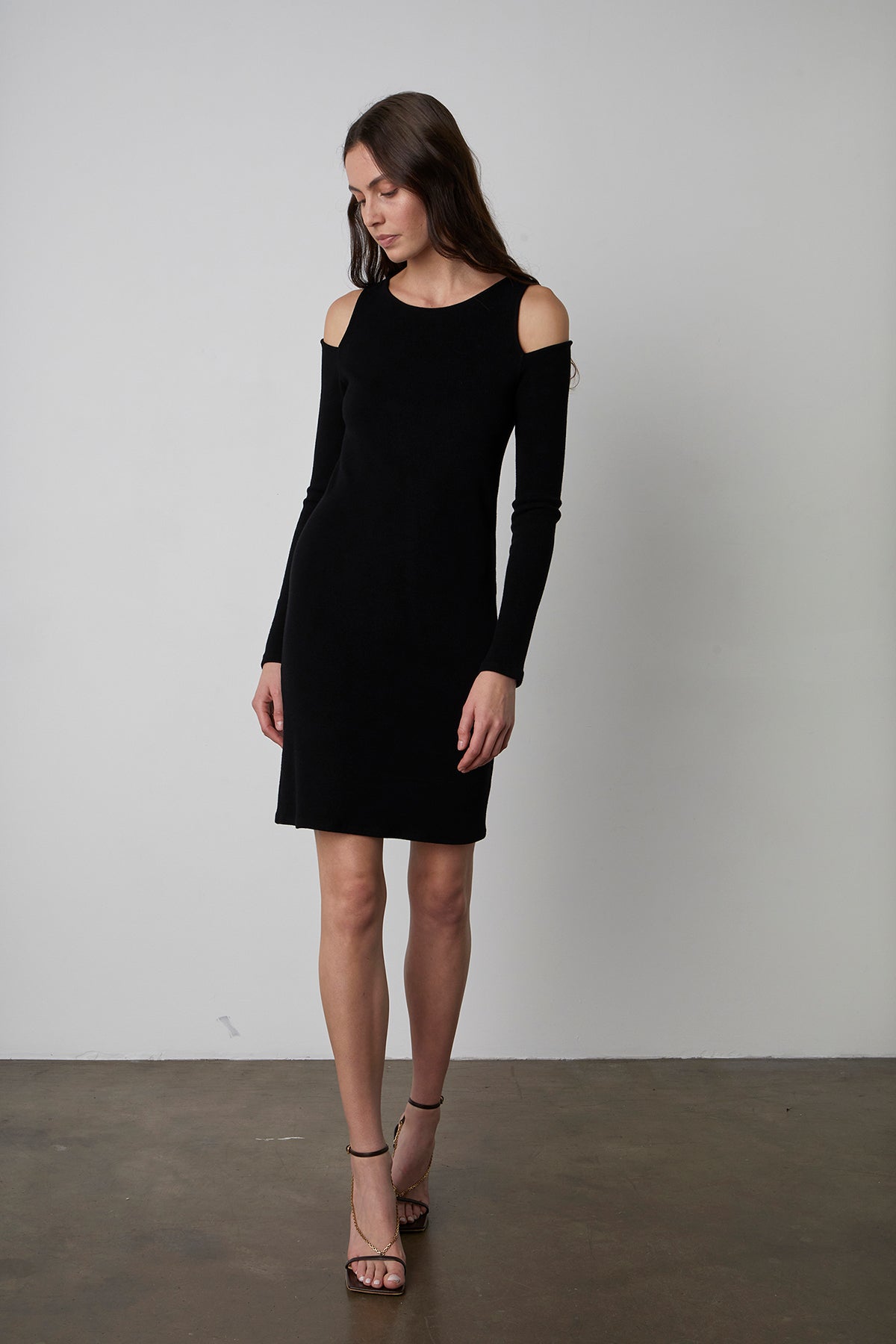 A woman stands against a plain background wearing a sleek LINNIE CUT-OUT DRESS by Velvet by Graham & Spencer with cold-shoulder details and strappy heels, looking down slightly.