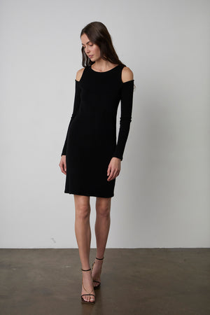 A woman stands against a plain background wearing a sleek LINNIE CUT-OUT DRESS by Velvet by Graham & Spencer with cold-shoulder details and strappy heels, looking down slightly.