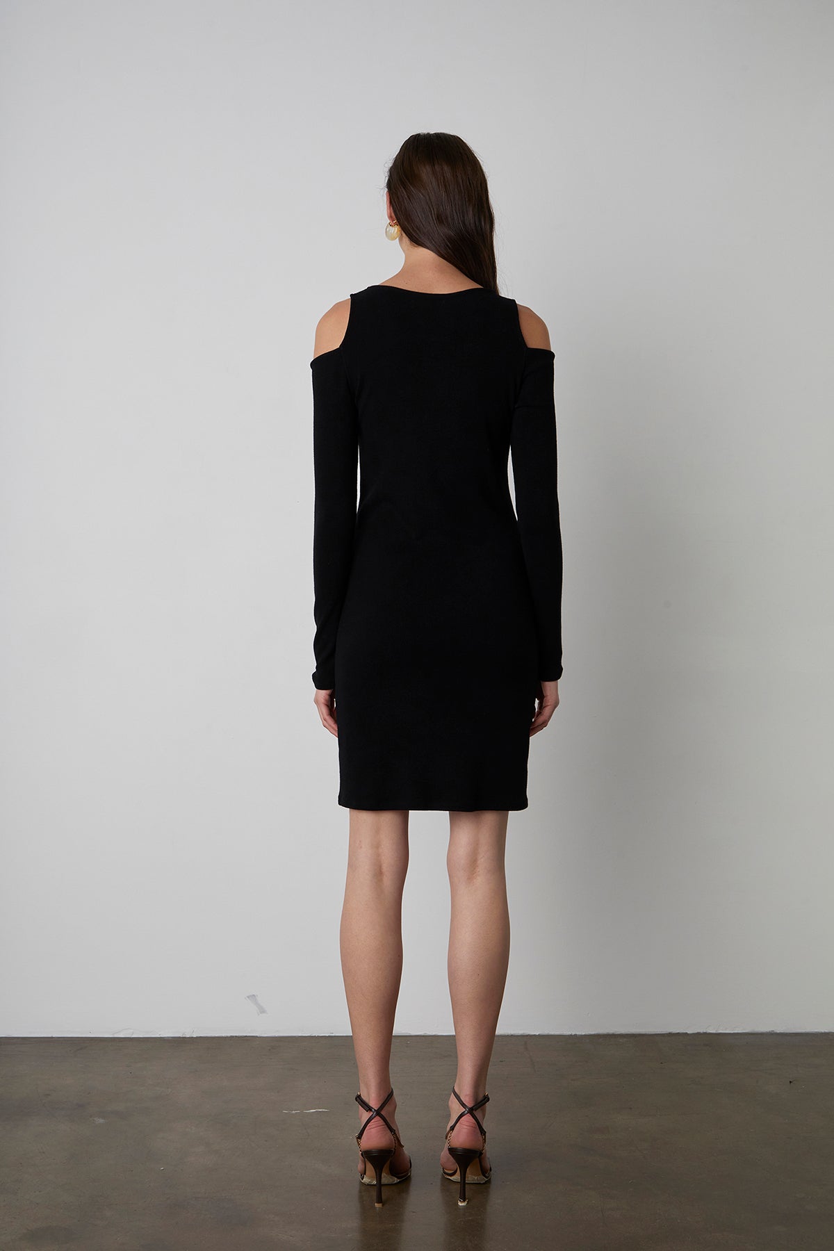 A woman stands facing away, wearing a black, long-sleeved, LINNIE CUT-OUT DRESS by Velvet by Graham & Spencer and strappy high heels. She has long, straight hair. The background is plain white.-23728333652161