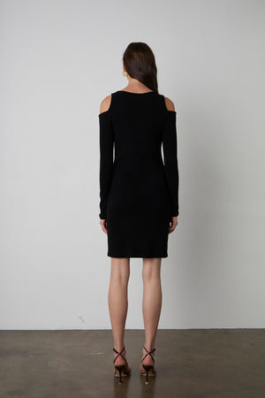 A woman stands facing away, wearing a black, long-sleeved, LINNIE CUT-OUT DRESS by Velvet by Graham & Spencer and strappy high heels. She has long, straight hair. The background is plain white.
