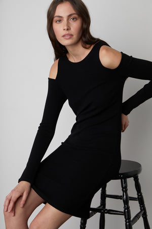 A woman with long brown hair is sitting on a black wooden stool. She is wearing the LINNIE CUT-OUT DRESS by Velvet by Graham & Spencer and looks at the camera.