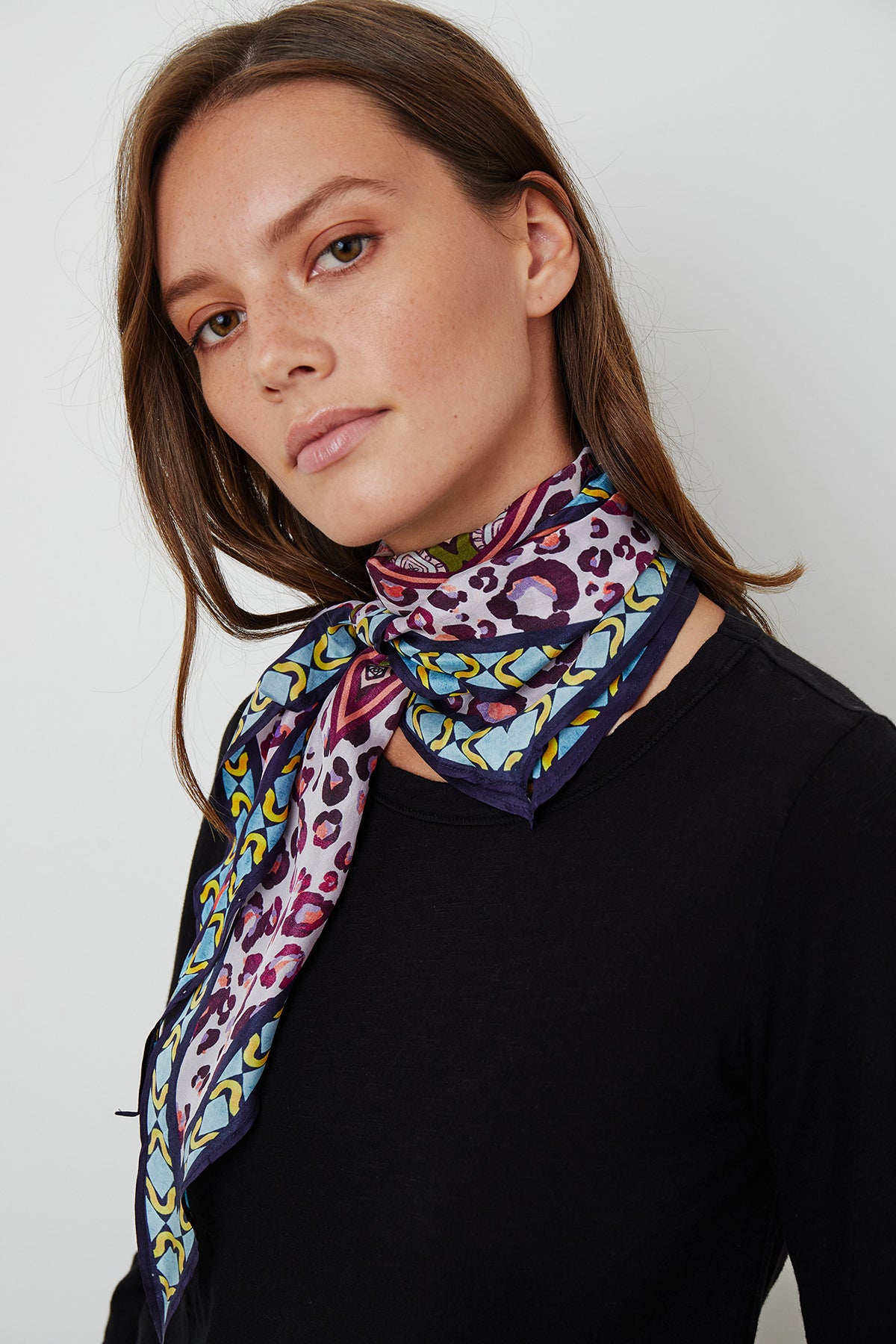 A woman with straight brown hair wearing a black shirt and a Velvet by Graham & Spencer DIAMOND SHAPE SILK SCARF loosely tied around her neck, standing against a plain white background.