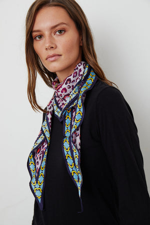 A person with long hair wears the vibrant DIAMOND SHAPE SILK SCARF from Velvet by Graham & Spencer around their neck and a black long-sleeve shirt, standing against a plain white background.