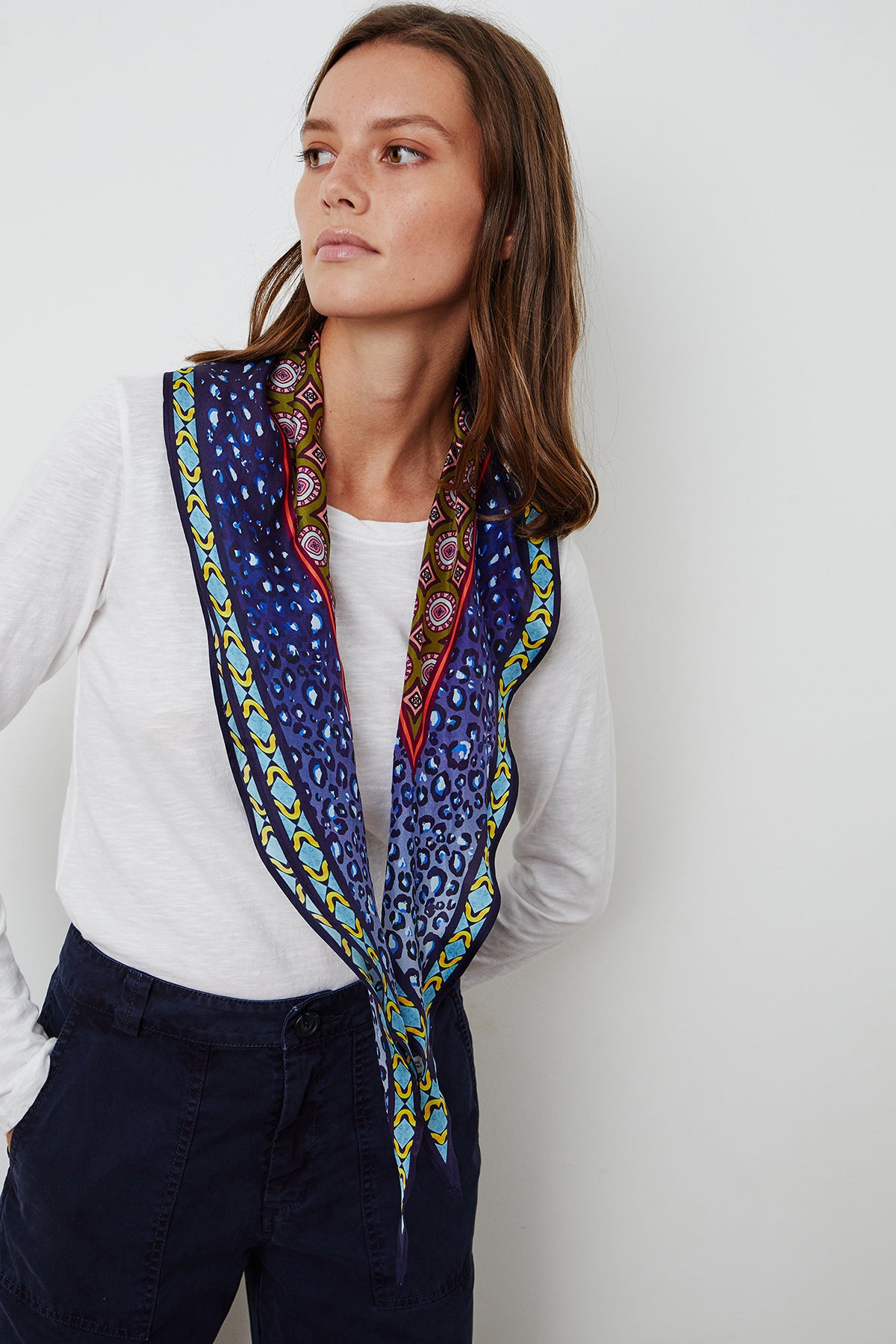 A person wearing a white long-sleeve shirt, dark pants, and the DIAMOND SHAPE SILK SCARF by Velvet by Graham & Spencer, featuring vibrant blue and yellow patterned designs, looks to the side against a plain white background.