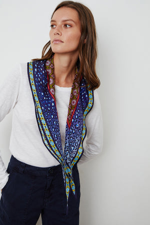 A person wearing a white long-sleeve shirt, dark pants, and the DIAMOND SHAPE SILK SCARF by Velvet by Graham & Spencer, featuring vibrant blue and yellow patterned designs, looks to the side against a plain white background.