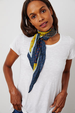 A woman with long hair wears a white t-shirt and the DIAMOND SHAPE SILK SCARF from Velvet by Graham & Spencer around her neck. She has her hands in her pockets and is looking directly at the camera.