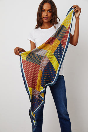 A person standing, holding a multicolored DIAMOND SHAPE SILK SCARF by Velvet by Graham & Spencer.