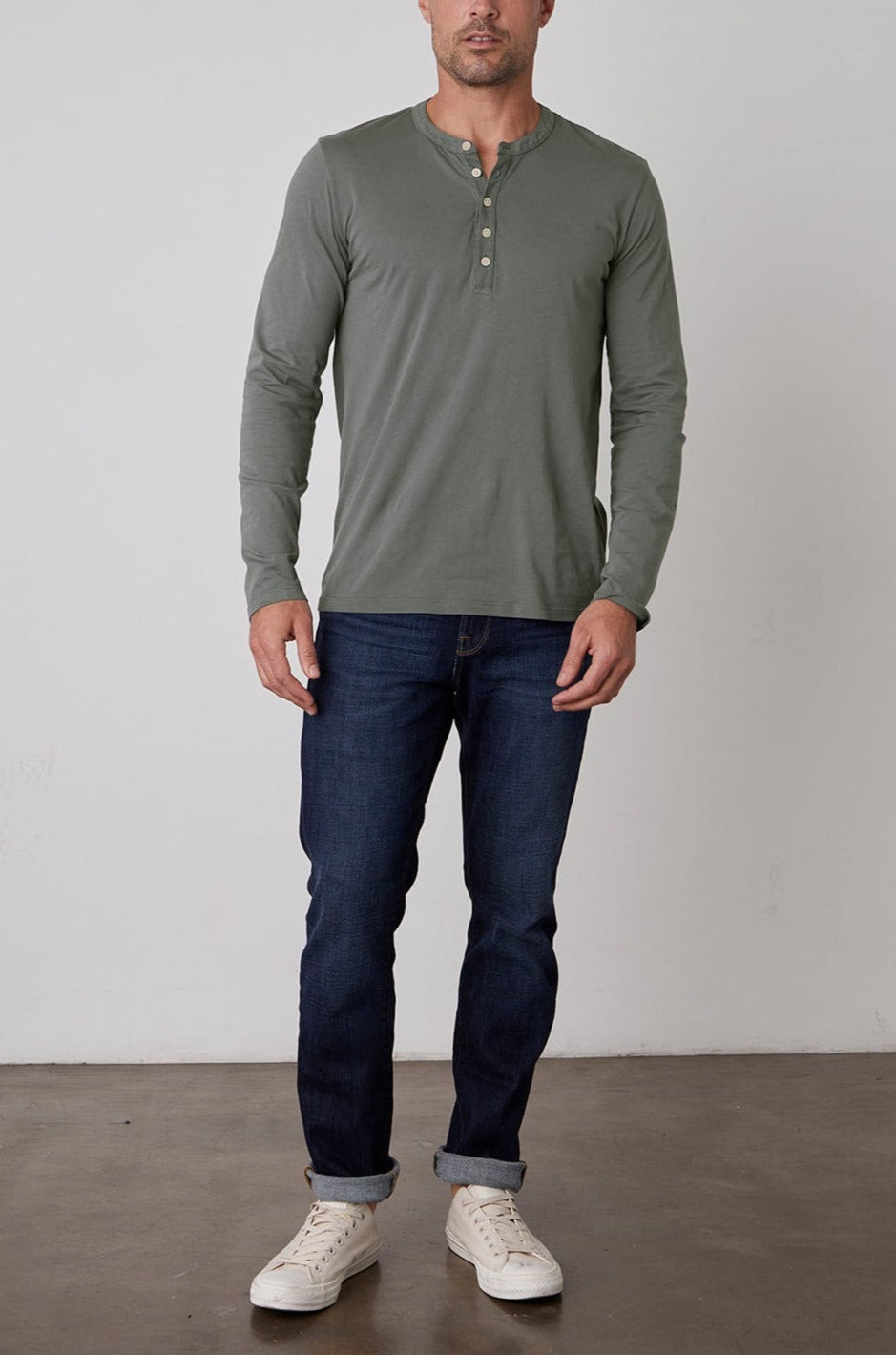 A man standing against a plain background is wearing a green ALVARO HENLEY by Velvet by Graham & Spencer, dark blue jeans, and white sneakers.-23778972467393