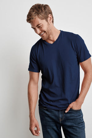A man with short brown hair and a beard is smiling while wearing a navy blue SAMSEN WHISPER CLASSIC V-NECK TEE by Velvet by Graham & Spencer, perfect for easy layering, and jeans. He has one hand in his pocket and is looking down.
