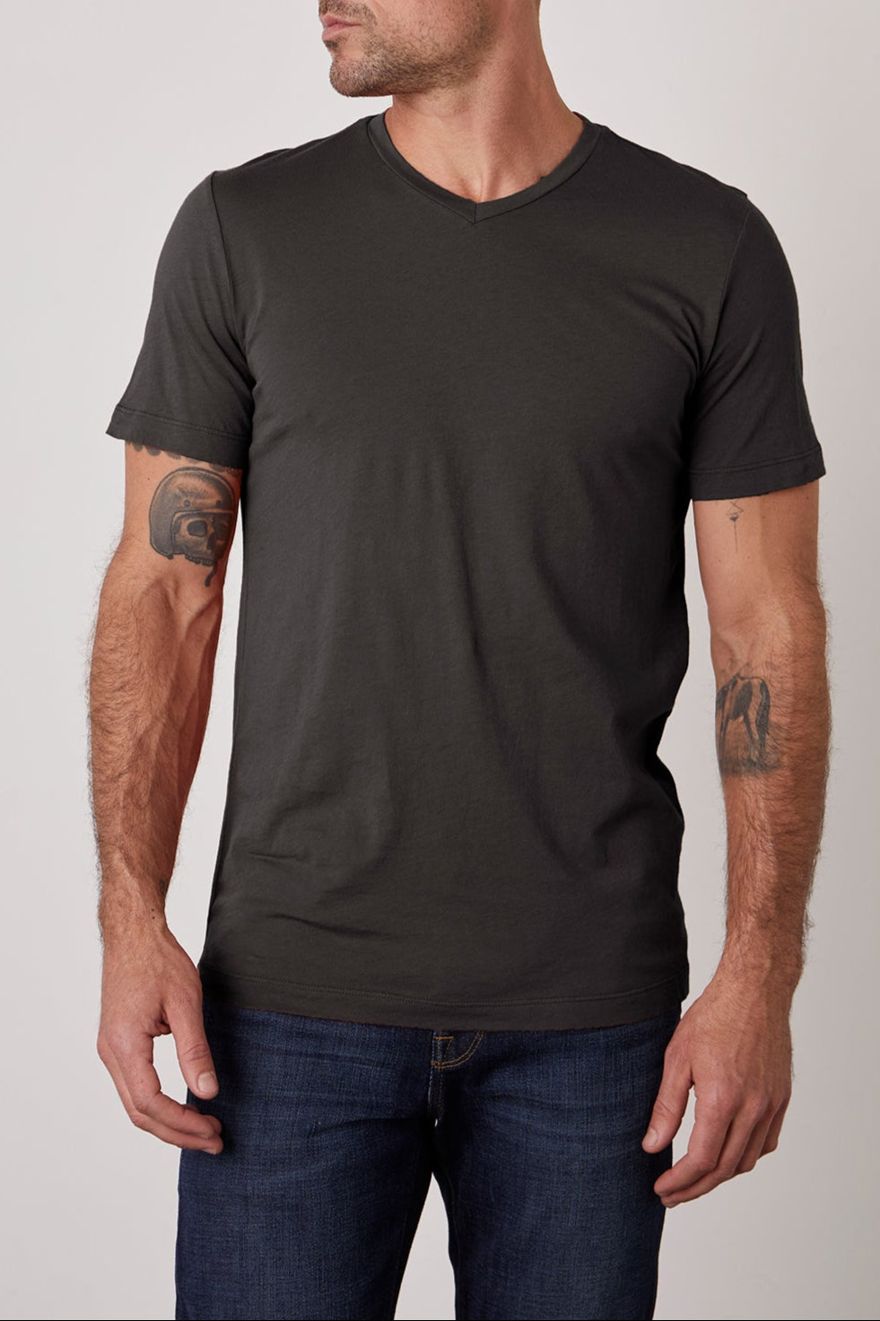 A man with a shaved head and tattoos on his forearms is wearing a plain, fitted black SAMSEN TEE by Velvet by Graham & Spencer and dark jeans. He is looking off to the side against a plain background.-25288803745985