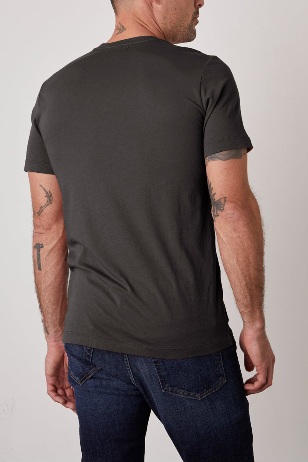 A man with a shaved head and tattoos on his neck and arms is standing and facing away, wearing a dark grey SAMSEN TEE by Velvet by Graham & Spencer that showcases a flawless fit and dark blue jeans.