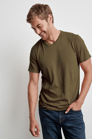 A man with short hair, wearing a green SAMSEN WHISPER CLASSIC V-NECK TEE by Velvet by Graham & Spencer and blue jeans, stands against a plain background with a hand in his pocket, looking down and smiling.