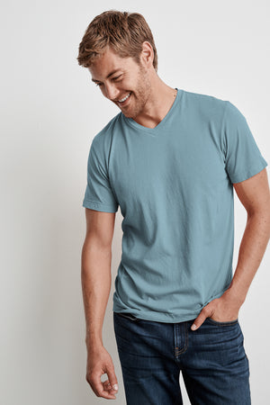 A man wearing a light blue Velvet by Graham & Spencer SAMSEN WHISPER CLASSIC V-NECK TEE with a whisper cotton knit and dark jeans stands against a plain white background, looking down and smiling. The easy layering of his outfit gives off a casual yet stylish vibe.