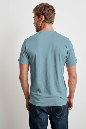 A man stands facing away from the camera, wearing a light blue SAMSEN WHISPER CLASSIC V-NECK TEE from Velvet by Graham & Spencer and dark blue jeans. The whisper cotton knit of his shirt ensures comfort and easy layering for any occasion.