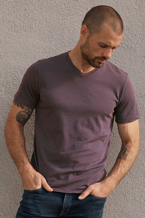 A man with a shaved head and tattoos on his arms, wearing a short-sleeved purple SAMSEN WHISPER CLASSIC V-NECK TEE by Velvet by Graham & Spencer and blue jeans, stands against a textured gray wall, looking down with his hands in his pockets.