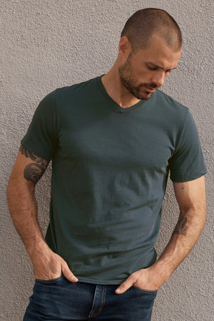A man with a shaved head and tattoos stands against a textured wall, wearing a dark green SAMSEN WHISPER CLASSIC V-NECK TEE from Velvet by Graham & Spencer and jeans, with his hands in his pockets.