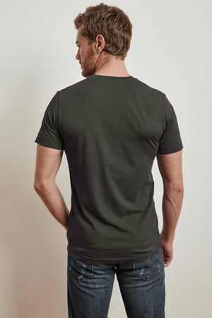 A man stands with his back to the camera, wearing a dark t-shirt and jeans. His SAMSEN WHISPER CLASSIC V-NECK TEE by Velvet by Graham & Spencer looks perfect for layering, hinting at the comfort of whisper cotton knit.