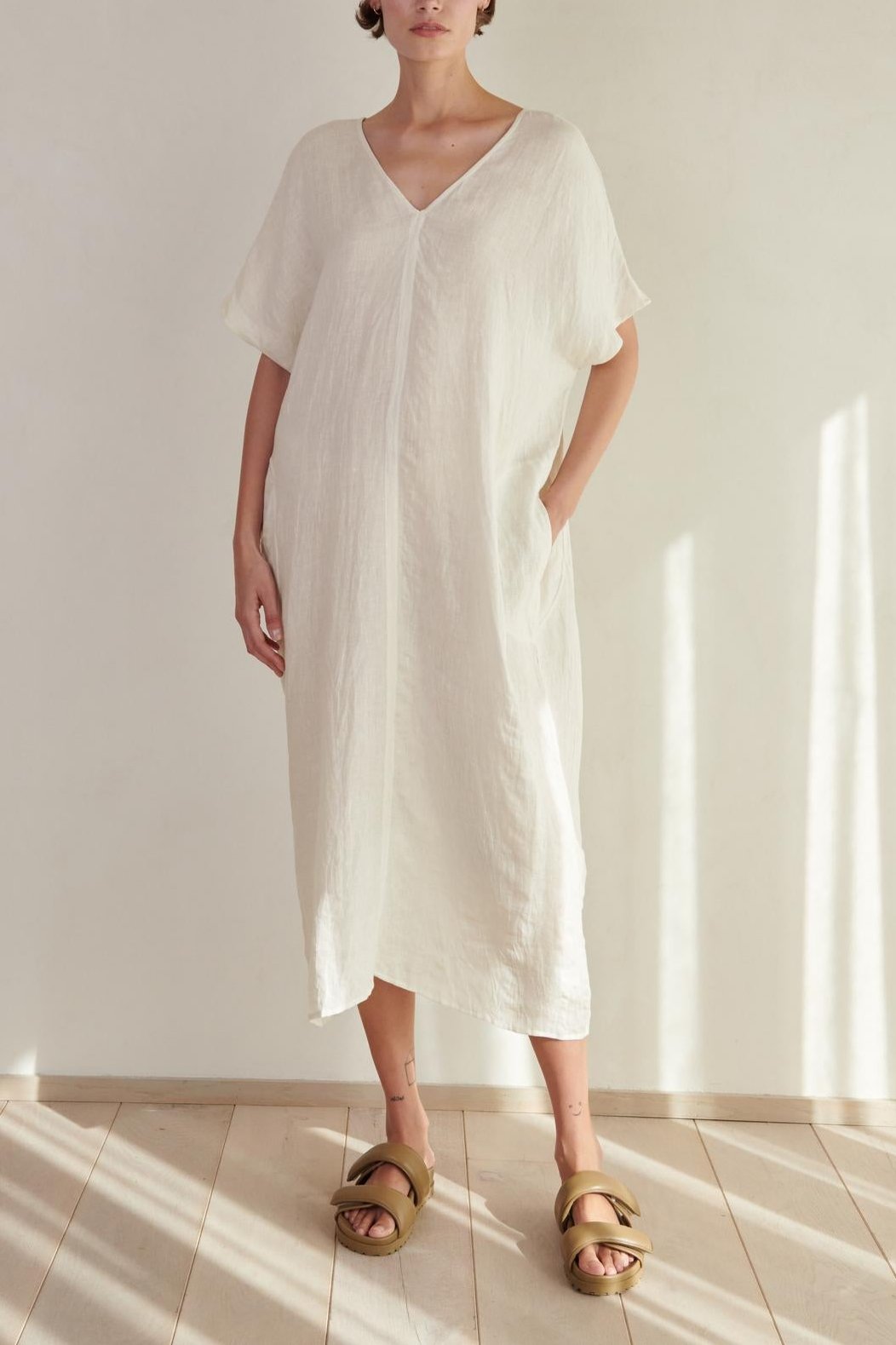 A woman wearing a long, white MONTANA LINEN DRESS with a V neckline and gold sandals stands in a softly lit room with a wooden floor. (Brand Name: Velvet by Jenny Graham)-26293194850497