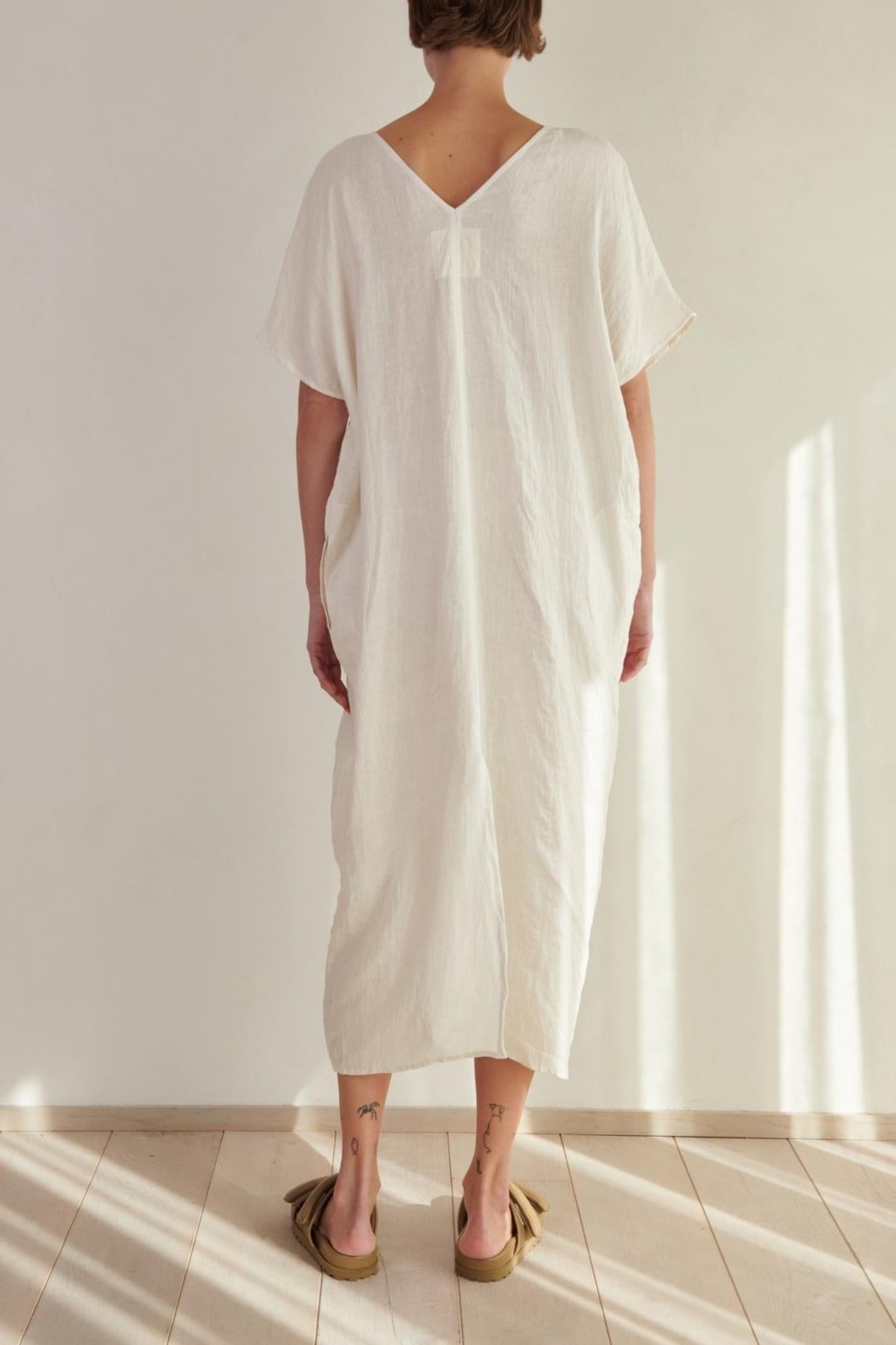 A woman wearing a Velvet by Jenny Graham Montana Linen Dress stands with her back to the camera in a bright, sunlit room.
