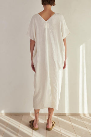 A woman wearing a Velvet by Jenny Graham Montana Linen Dress stands with her back to the camera in a bright, sunlit room.