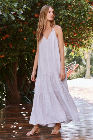 A woman stands outdoors wearing the CLEO LINEN MAXI DRESS by Velvet by Graham & Spencer, a light gray maxi dress with spaghetti straps. She is under an orange tree and there are more trees and some furniture in the background.