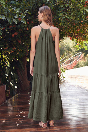 A woman stands on a wooden deck facing away, wearing the Velvet by Graham & Spencer CLEO LINEN MAXI DRESS in green and sandals, with greenery and outdoor furniture in the background.