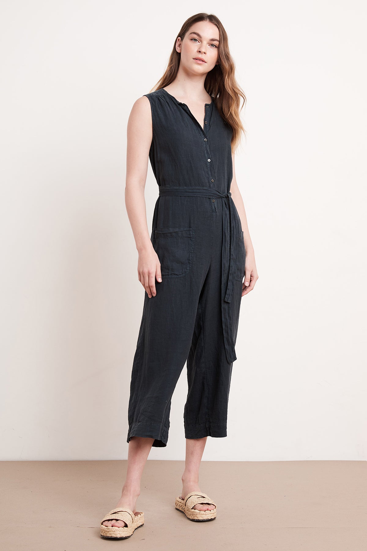A person stands indoors against a white background, wearing the DANI LINEN JUMPSUIT by Velvet by Graham & Spencer with a tied waist and sandals. The relaxed silhouette of this sleeveless black jumpsuit, crafted from medium-weight linen, offers an easy breezy style perfect for any casual occasion.