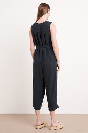 A woman, with long hair flowing down her back, stands facing away while wearing the DANI LINEN JUMPSUIT from Velvet by Graham & Spencer. This sleeveless black jumpsuit, designed with a tied waist and crafted from medium-weight linen, boasts a relaxed silhouette. She pairs her look with beige flat sandals against a plain white background.