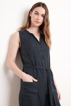 A woman with long hair stands against a white background, wearing the DANI LINEN JUMPSUIT by Velvet by Graham & Spencer, featuring a sleeveless design with large front pockets and a belt, showcasing a relaxed silhouette.