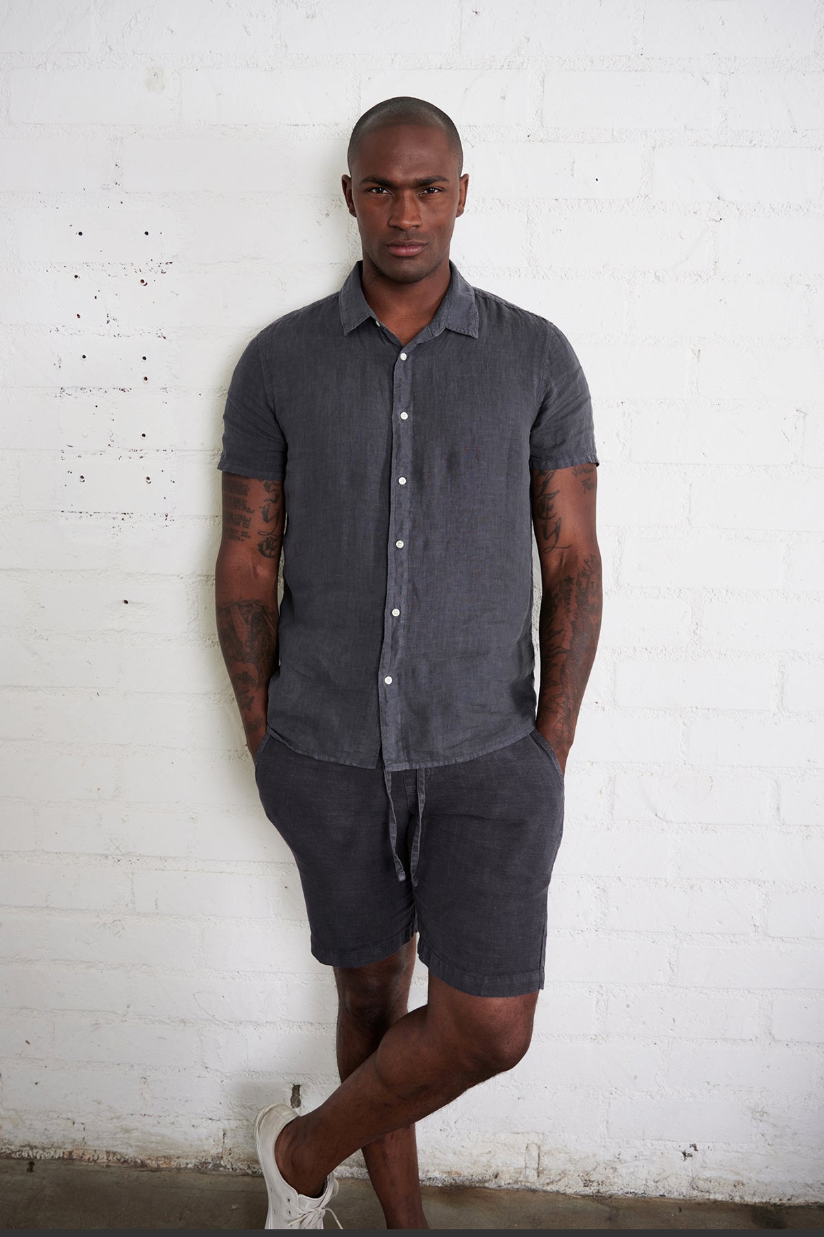 Mackie linen button up shirt in carbon with model standing against white brick wall with hands in pockets.